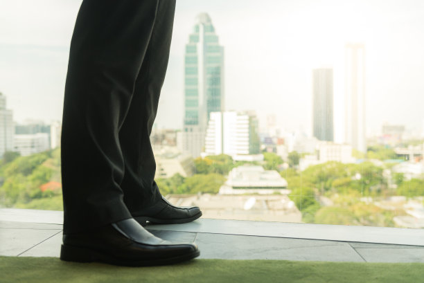 全长后视图的成功商人西装站在办公室与手在他的腰，首席执行官看着透过窗户在大城市的建筑，规划新的项目，等待会议开始图片下载