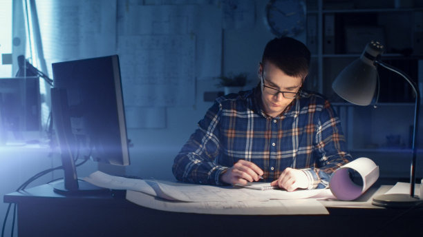 年轻能干的建筑工程师晚上在他的办公室工作。办公室很暗，只有桌上的灯是亮着的。摆在桌上的各种计划和蓝图。他在记事本上画画。图片下载