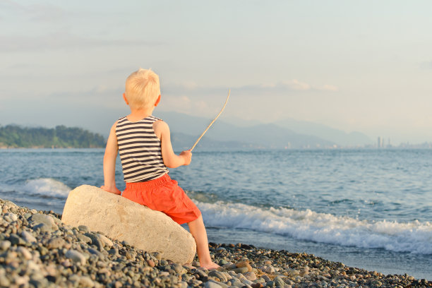 穿着条纹t恤的男孩拿着棍子坐在沙滩上。后视图图片下载