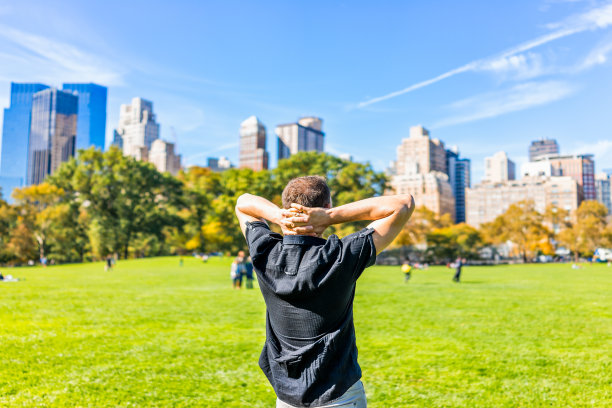 一个快乐的年轻人双手抱头，站在纽约中央公园在阳光明媚的秋日摩天大楼和人图片下载