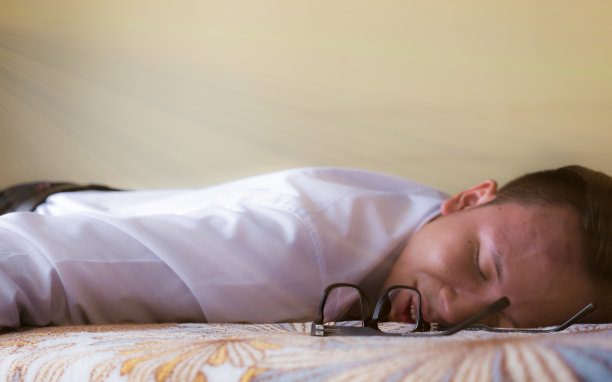 一个疲惫不堪的年轻商人戴着眼镜躺在床上，在旅行后睡着了，柔和的色调带着淡淡的光线图片下载