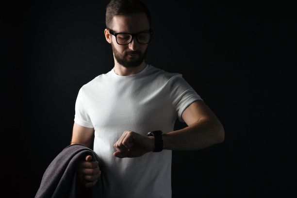 一名戴着透明眼镜、身穿白色空白t恤、蓄着胡须的年轻男子正在看智能手表，手里拿着一件外套图片下载