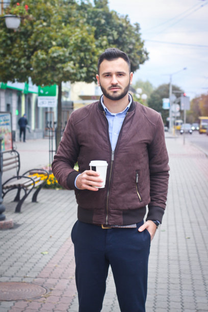 一个穿着夹克和西装的男人走在欧洲的大街上，手里拿着咖啡。商人时尚服装。秋天的风景、肖像。库存图片图片下载