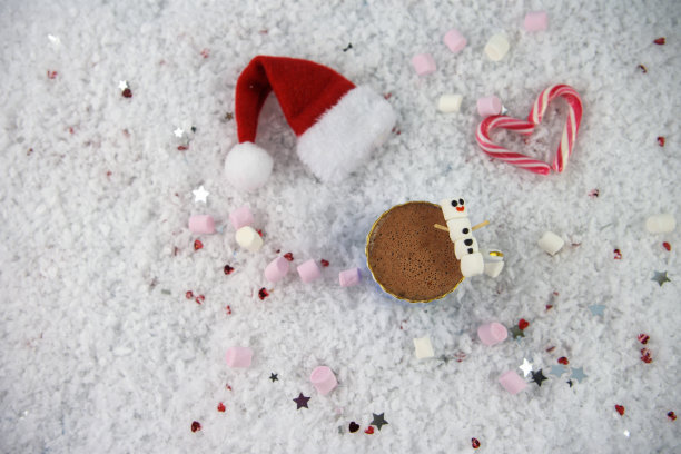 圣诞食物和饮料的图片，一杯热巧克力和迷你棉花糖的形状为快乐的雪人与冰微笑放在雪与红色圣诞帽，撒有小心和星星图片下载