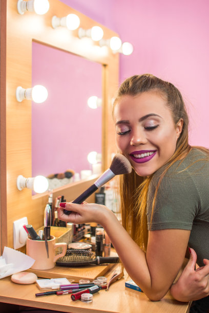 美丽微笑的女孩靠在梳妆台上，拿着镜子在擦粉。图片下载