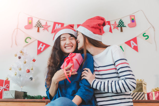 年轻美丽的亚洲女人赠送礼盒和亲吻最好的女性朋友。笑脸室以圣诞树装饰为节日背景。圣诞派对和庆祝概念。素材图片