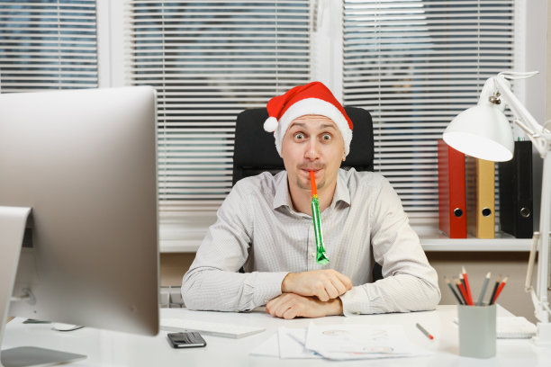 英俊震惊的商人在衬衫和红色圣诞帽坐在桌子上，工作在电脑与现代显示器，灯，文件和生日烟斗在灯光办公室的窗户背景。图片下载