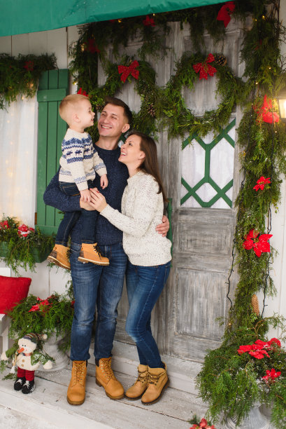 快乐的父母和小儿子。小男孩穿着毛衣呆在门廊雪台阶上的灯塔与装饰在家里的红绿新年门。圣诞好心情。家庭和假日2018概念图片下载