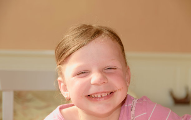 一个可爱的小女孩微笑着。一个脸上有巧克力奶油污渍的小女孩。一个小女孩坐在厨房里。图片下载