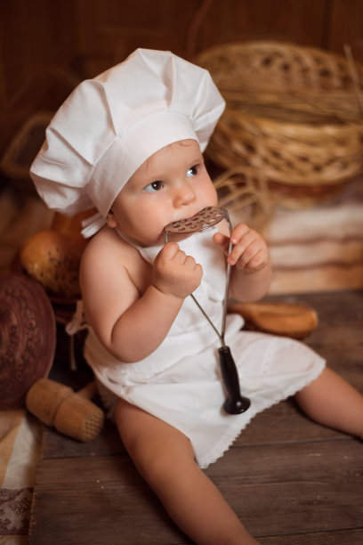 快乐的小男孩穿着厨师服坐在厨房的桌子上，旁边的篮子里装着美味的面包卷、小圆面包和面包。本空间图片下载