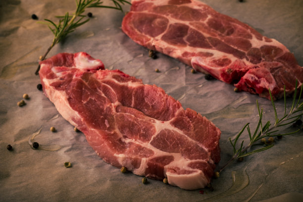 两片猪颈牛排的横向照片。红色和脂肪的肉部分与胡椒，迷迭香树枝放在破旧的烤纸上。到处都是飞溅的油。图片下载