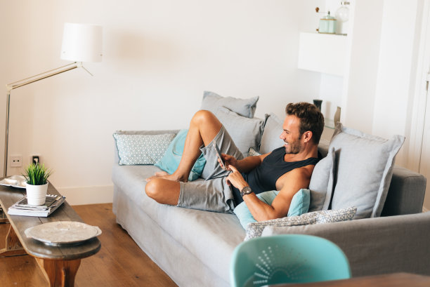 快乐年轻的金发男子坐在家里的沙发上看手机图片下载