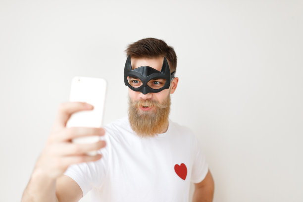 孤立的照片，有吸引力的男性与厚厚的长胡子和胡子覆盖脸蝙蝠面具，看着智能手机，摆姿势自拍，穿着休闲的白色t恤。超级英雄模特在室内用手机图片下载