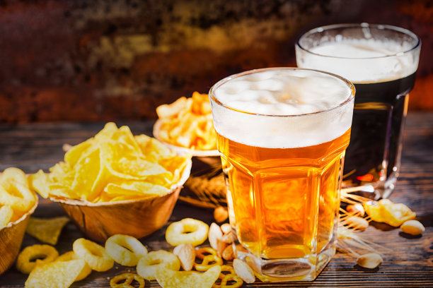 深色木桌上放着两杯淡啤和黑啤，上面有大块泡沫，盘子旁边放着薯条和零零碎碎的零食图片下载