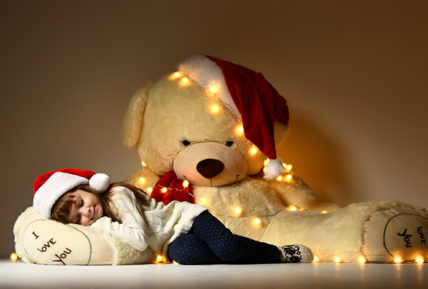年轻女孩睡觉与大的软泰迪熊玩具在圣诞老人的红帽子图片下载