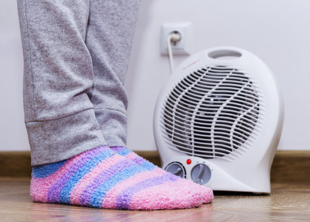 一个穿着暖冬袜子的女人在热风扇旁暖脚。家里有电暖器。图片下载