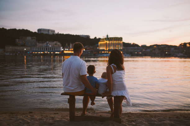 概念家庭海滩度假。一个四口之家，妈妈，爸爸，兄弟，孩子，男孩在夏天日落时坐在河边的木凳上图片下载