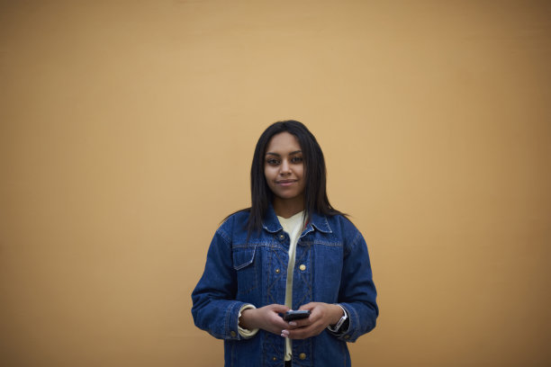 肖像迷人的黑色模特穿着时尚的牛仔裤夹克发送照片给朋友站在墙上宣传背景，积极的国际女学生通过智能手机邮寄图片下载