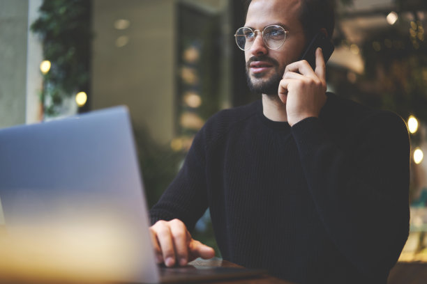 照片中，一位戴着眼镜的男性经理坐在咖啡厅里通过智能手机与客户交谈，并使用可以连接无线互联网的笔记本电脑进行远程工作图片下载