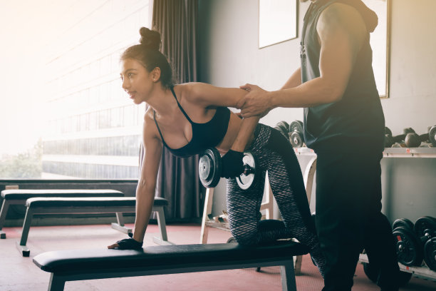 在室内健身房，男士帮助女士举起哑铃进行个人训练。图片下载