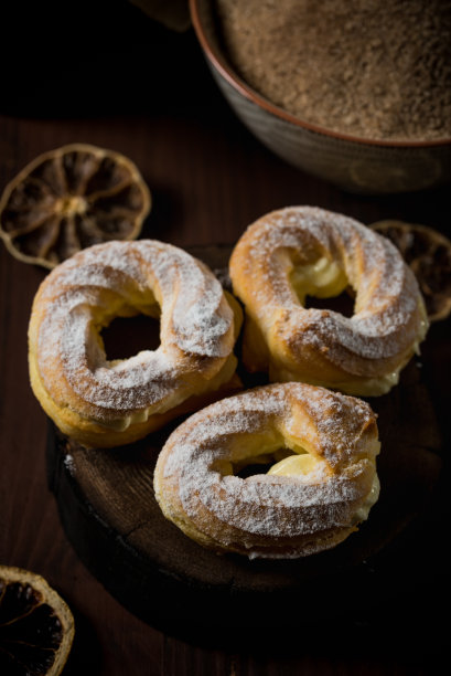 垂直照片与三个自制泡芙塞满甜奶油。这些泡芙上面覆盖着糖粉。黑色的木板上散落着一些干橘色的圆环。图片下载