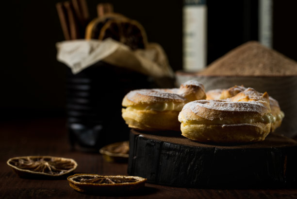 水平照片与三个自制泡芙塞满甜奶油，葡萄酒和肉桂棒。这些泡芙上面覆盖着糖粉。黑色的木板上散落着一些干橘色的圆环。图片下载