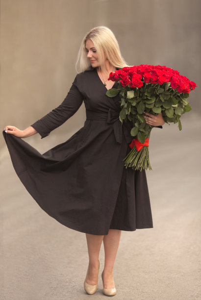 美丽的金发女人的肖像，穿着黑色复古的连衣裙，手持一百朵红玫瑰的大花束户外。惊喜礼物在日期。图片下载