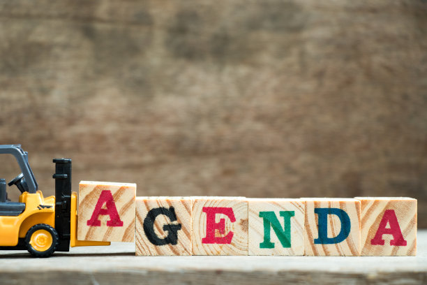 黄色玩具叉车保持字母块A，以完成单词议程在木材背景上图片下载