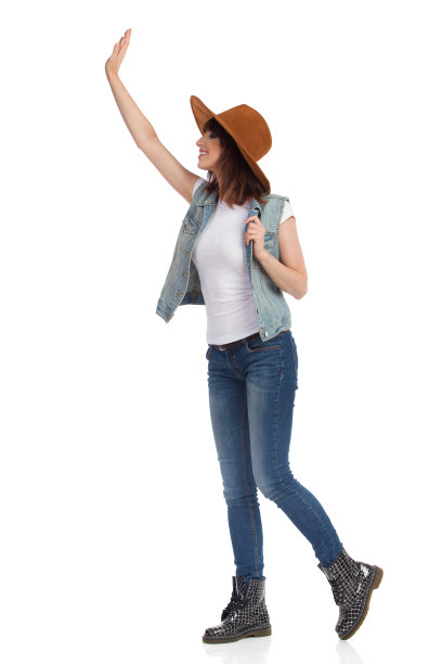 年轻女子在棕色帽子，牛仔裤背心和靴子是站和挥手。侧视图。图片下载