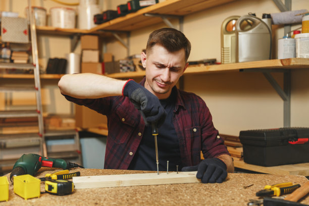 英俊微笑的白人青年男子在格子衬衫，黑色t恤，手套拧螺丝，在木工车间的木桌地方与一块木头，不同的工具。图片下载