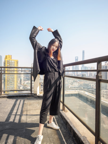 身着黑色外套的黑发美女站在大厦屋顶上，在模糊的外滩地标建筑背景中伸展双臂。情感、人、美、生活理念。图片下载