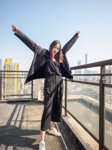 身着黑色外套的黑发美女站在大厦屋顶上，在模糊的外滩地标建筑背景中伸展双臂。情感、人、美、生活理念。图片下载