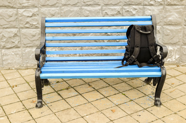 公园里铺着瓷砖的人行道上的蓝色长椅，背着黑色的背包。没有身体。图片下载