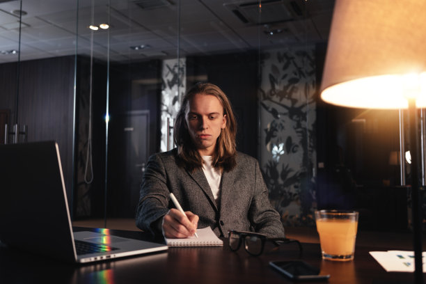 疲惫的年轻商人长头发在晚上工作现代联合工作室。男人坐在木桌旁，使用现代笔记本和办公工具图片下载