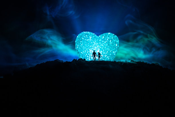 夜晚，一对幸福的夫妇站在山的大形状的心符号后面的剪影。巨大的心像月亮在朦胧的天空发光。情人节装饰照片图片下载