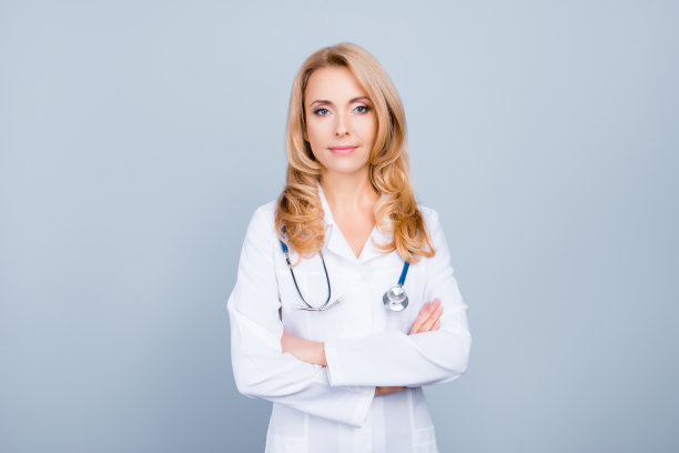 有魅力的女医生穿着白色制服，脖子上挂着听诊器，双手交叉，灰色背景，表情严肃素材图片