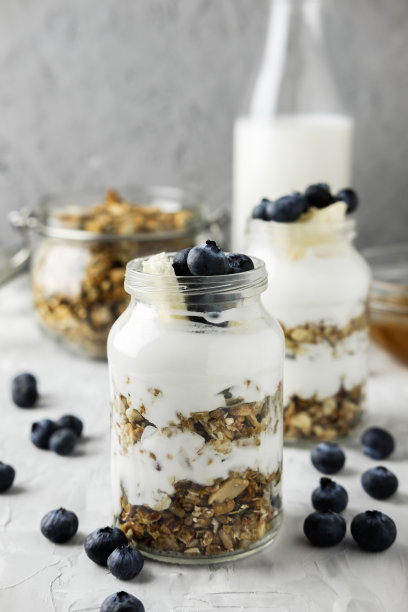 自制的格兰诺拉麦片装在一个玻璃罐里，里面有酸奶和蓝莓，背景是灰色的混凝土，还有一瓶牛奶和蜂蜜。健康早餐的食物照片。图片下载