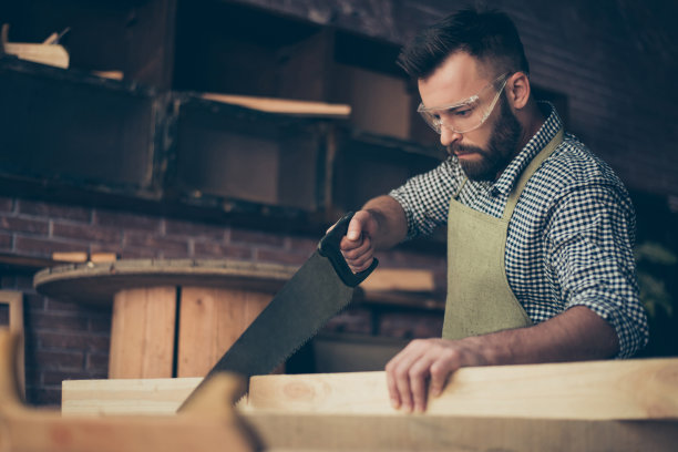近距离的照片自信英俊的胡子熟练穿着围裙格子衬衫和保护眼镜木匠，他正在用一个锯子把木板分成两部分图片下载