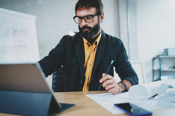 照片中，一名留着胡子的年轻男性建筑师戴着眼镜，在办公桌前的数字平板电脑基座上工作。专业经验丰富的施工工程师，水平，模糊背景。图片下载