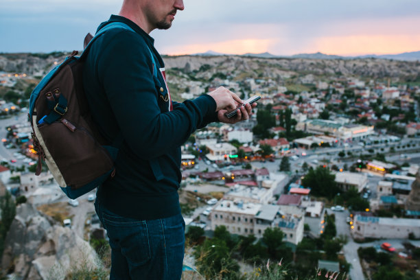 一名游客在夕阳的背景下背着背包，看着手机上的地理定位和移动应用程序，准备打电话。背景是傍晚的夕阳和城市。图片下载