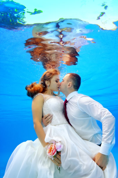 在阳光的背景下，美丽的男人和女人在游泳池里穿着婚纱拥抱和亲吻。肖像。在水下拍摄。垂直方向图片下载