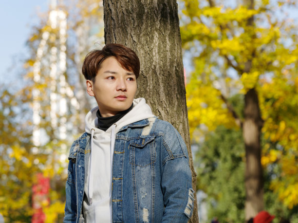 年轻的中国微笑男子肖像坐在模糊的黄色树背景在日落在秋天公园。户外。图片下载