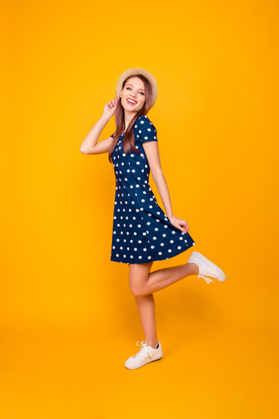 全长，全身肖像的漂亮，愉快，积极，可爱的女孩与抬高的腿，手在帽子和波尔卡点连衣裙底部，摆姿势在黄色的背景图片下载