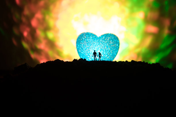 夜晚，一对幸福的夫妇站在山的大形状的心符号后面的剪影。巨大的心像月亮在朦胧的天空发光。情人节装饰照片图片下载
