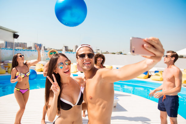 和平!快乐可爱的情侣摆姿势自拍，小伙子拍照，回忆，假期，美好的晴天，他们和朋友在海滩附近的派对，他们玩球，带着巨大的缎带蓝色气球图片下载