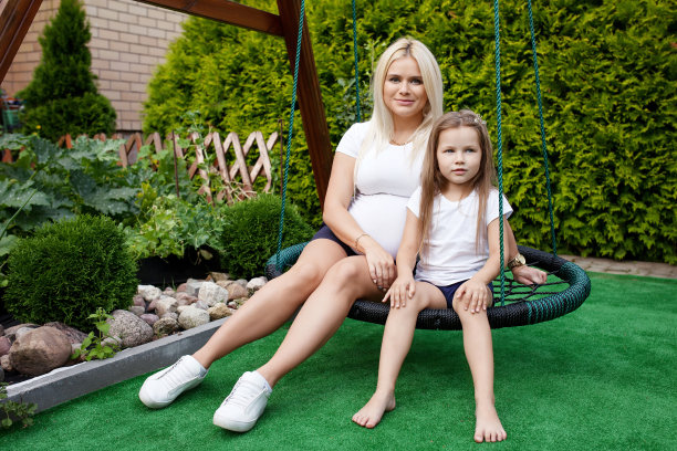 漂亮的孕妇和小女儿。可爱的小女孩和她怀孕的妈妈坐在夏日的秋千上。图片下载