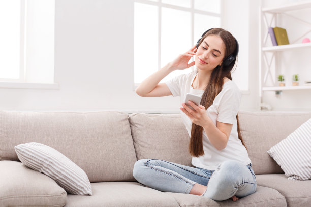 一名年轻女子坐在米色沙发上，拿着手机和耳机图片下载