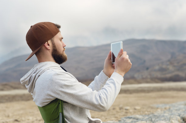 一个戴着胡子戴着帽子、穿着运动衫、手里拿着平板电脑的嬉皮士，在湖边的自然环境中用平板电脑拍照图片下载