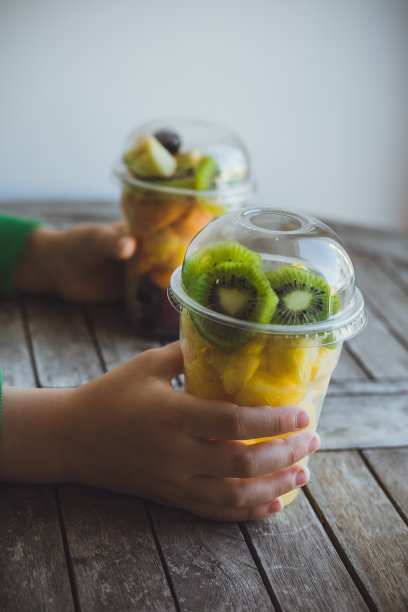 垂直照片男孩手拿两个塑料杯切水果:猕猴桃，菠萝，橘子，苹果，甜瓜图片下载