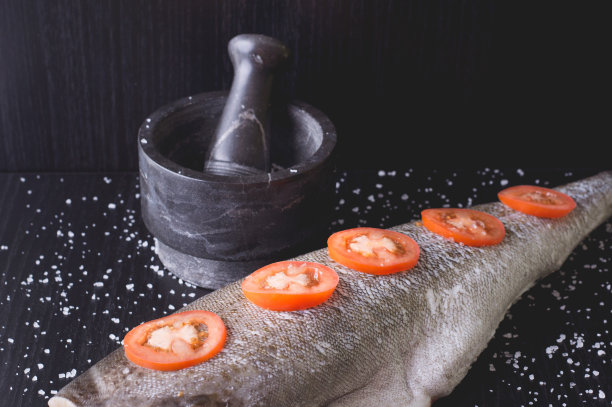 白色的鱼，西红柿和大理石砂浆在黑色的木制背景。烹饪的过程图片下载